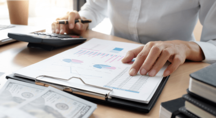 man's fingers are kept on a graph sheet which is attached with a notepad and another hand kept a pen and busy with a calculator