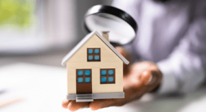 A man is holding a microscope in one hand and a small toy of house in other hand zooming microscope above the small house.